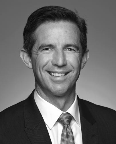 Black and white head shot of Simon Birmingham, former Minister for Finance. He is smiling and wearing a suit, shirt and tie.