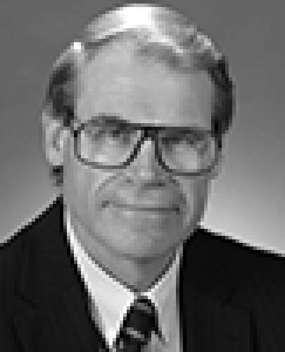 Black and white head shot of John Faulkner, former Special Minister of State. He is smiling and wearing a dark suit jacket, white shirt, tie, and large glasses.