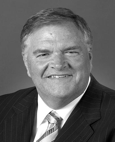 A black and white head shot of Kim Beazley, who is smiling and wearing a dark jacket, shirt and tie. 