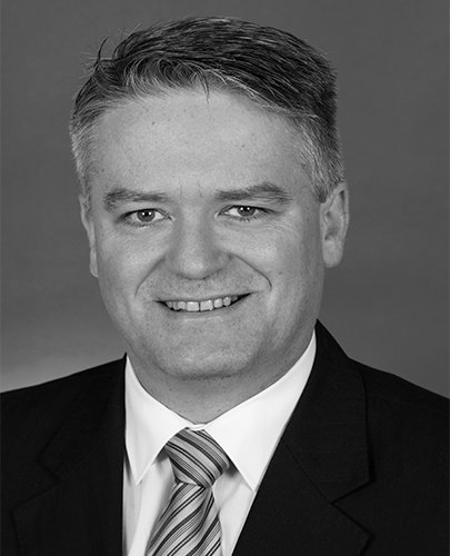 A black and white head shot of Mathias Cormann, who is smiling and wearing a dark jacket, shirt and tie. 