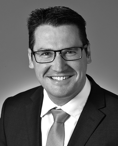 A black and white head shot of Zed Seselja, who is smiling and wearing a dark jacket, shirt, and tie. 