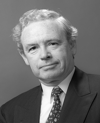 A black and white head shot of Ralph Willis, who is smiling and wearing a dark jacket, shirt and tie. 