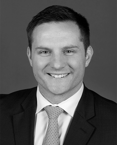 A black and white head shot of Alex Hawke, who is smiling and wearing a dark jacket, shirt, and tie. 