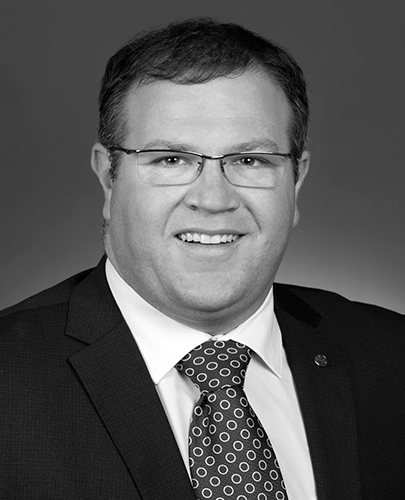 A black and white head shot of Ben Morton, who is smiling and wearing a dark suit, shirt and tie. 