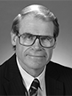 Black and white head shot of John Faulkner, former Special Minister of State. He is smiling and wearing a dark suit jacket, white shirt, tie, and large glasses. 