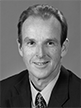 Black and white head shot of Joe Ludwig, former Special Minister of State. He is smiling and wearing a dark suit jacket, shirt, and tie. 