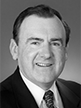 Black and white head shot of Dr David Kemp, former Minister Assisting the Minister for Finance for Privatisation. He is smiling and is wearing a dark suit jacket, white shirt and a tie. 