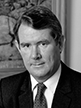 Black and white head shot of Peter Walsh, former Minister for Finance. He is smiling and wearing a dark jacket, shirt and tie. 