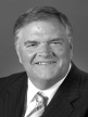 A black and white head shot of Kim Beazley, who is smiling and wearing a dark jacket, shirt and tie. 