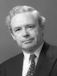 A black and white head shot of Ralph Willis, who is smiling and wearing a dark jacket, shirt and tie. 