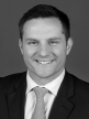 A black and white head shot of Alex Hawke, who is smiling and wearing a dark jacket, shirt, and tie. 